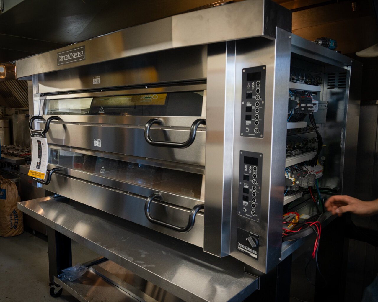 The brand‑new PizzaMaster Model 942 double‑deck electric pizza oven during installation, showing its stainless‑steel body, dual glass‑front baking chambers, heavy insulated doors, digital control panels, and open side access revealing the internal wiring as technicians hook it up — the same oven now used to fire every Back of House pizza at The Lancaster Smokehouse.
